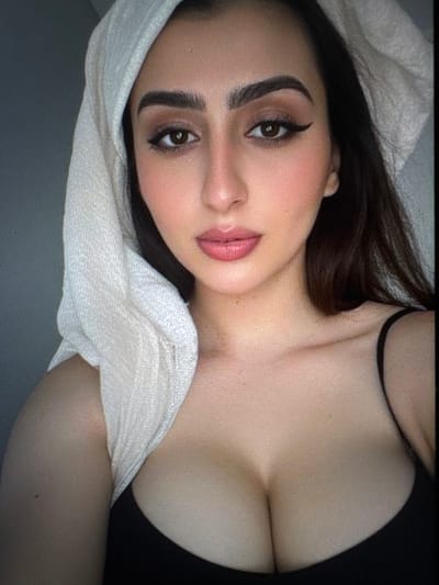 A woman lies with her head resting on a white woven cloth. Her dark hair frames a face with strong brows, lined eyes, and full lips. She wears a dark top with thin straps, revealing part of her chest. The image is taken from above, accentuating her features and the curves of her body.
