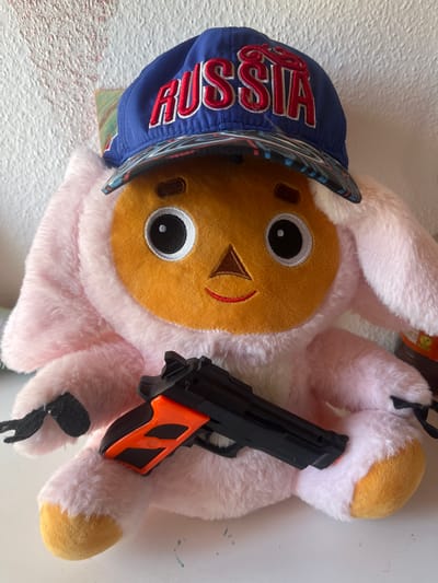 The image shows a plush toy, resembling a pink creature with a round orange face and large eyes, wearing a blue baseball cap with "RUSSIA" embroidered in red. The toy is holding a black and orange toy gun. The background consists of a textured white wall, with some faint red markings. A glimpse of a colorful painting or artwork is visible on the left side of the image. The toy is sitting on a white surface. No specific location in Schwäbisch Hall, Germany is identifiable from the image.