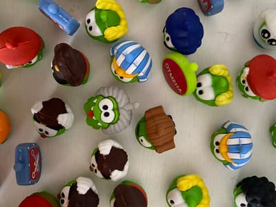 The image is a top-down, close-up shot of a collection of small, brightly colored toy figurines scattered on a white, textured surface. The figurines appear to be character toys, many of which are green with large eyes and some have accessories like hats or stripes. Some are wearing red hats, some are wearing brown hats, and one is wearing a blue, striped outfit. Another figurine has a red cap with white lettering that reads "ОТМЕНА". There is also a blue toy that resembles a digital alarm clock. The arrangement is random, with some figurines overlapping. The lighting is even, casting minimal shadows. There is no indication of any specific location in Ostrovki, Russia, or any human presence in the image.