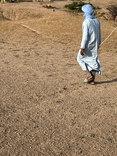 A man wearing a blue turban and a light blue garment stands in a dry, arid environment. The background suggests a location near the city of Sidi Badhaj, Morocco, based on the arid landscape. The ground is a mix of gravel and dry earth. A rope is visible. The person is wearing sandals or slip-on shoes.