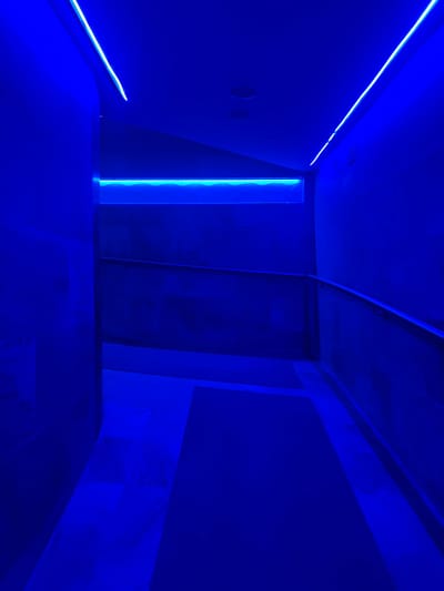 This image captures an indoor hallway or ramp structure, located in Bağcılar, Turkey, intensely illuminated by blue LED lighting. No individuals or specific actions are depicted. The ceiling features two linear blue LED strips running longitudinally, along with two circular recessed light fixtures. A horizontal blue LED strip, displaying a wavy light pattern, is recessed into the upper section of the left wall. The walls appear uniformly dark blue due to the pervasive lighting. On the right, a silver-toned handrail descends diagonally, indicating a downward slope or ramp that forms the primary floor surface. The floor itself is a dark, uniform material, reflecting the blue light. The environment presents as a modern, controlled, and distinctively lit architectural pathway.