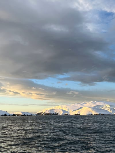 The image shows a landscape divided into three distinct sections. On the left, a cloudy sky filled with various shades of grey and some blue patches. The middle section features snow-capped mountains with golden sunlight. On the right is the sea, with visibly strong currents running through it.