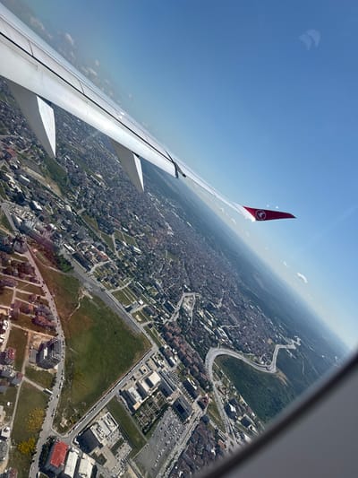 The image is an aerial view of a city, likely Bağcılar, Turkey, taken from an airplane window. The foreground is dominated by the white wing of the aircraft, angled across the frame, with visible flaps and supports. The wing has a red tip adorned with a white logo, which appears to be the Turkish Airlines logo. Below the wing, the city unfolds with a dense collection of buildings, characterized by a mix of residential structures with red and grey roofs, and larger, modern institutional buildings with flat, white roofs, some of which are surrounded by parking lots. A winding highway cuts through the urban landscape, bordered by green spaces and trees. In the distance, the urban sprawl continues, gradually fading into a hazy horizon. The sky is a clear, bright blue with a few scattered white clouds. The image is tilted, giving a dynamic perspective of the cityscape from above.