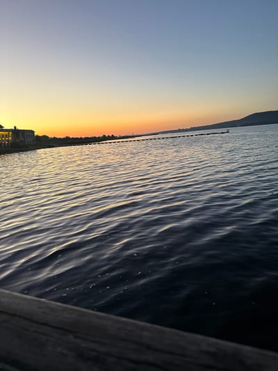 The image depicts a serene body of water at dusk, with a clear sky transitioning from a pale blue to a warm orange hue near the horizon. In the distance, a line of trees and structures is visible along the shoreline, and a long pier or breakwater extends into the water. A faint outline of a hill or mountain is present on the right side of the horizon. The foreground is dominated by the dark, rippling surface of the water, with reflections of the sunset catching the crests of the waves. A wooden railing or edge of a dock is visible in the lower left corner, partially obscuring the water. The overall atmosphere is peaceful and tranquil, suggestive of a quiet evening by the water. The specific location of Казашко, Bulgaria, is not visually identifiable from the image itself.