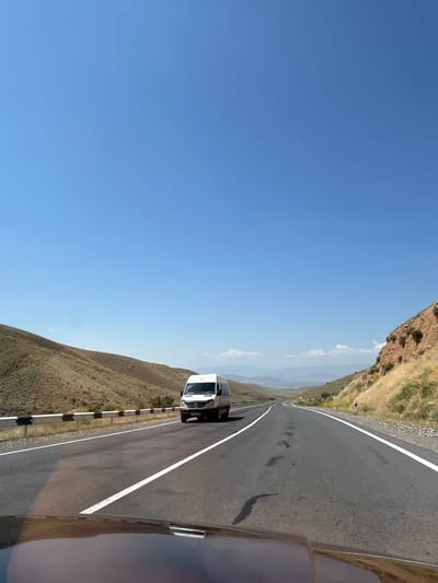 This media file depicts a view from a vehicle traveling on a paved road through a mountainous, arid landscape. The road is bordered by a steep, dry hillside on the left and a guardrail. The sky is a clear, bright blue, suggesting a sunny day. In the distance, a valley can be seen, with hazy mountains on the horizon.
The primary visible object is a white van driving on the road ahead. The perspective suggests the image was taken from another vehicle, possibly in the passenger seat or from a dashcam. The time of day appears to be daytime, likely midday or afternoon, given the bright sunlight.
The scene conveys a sense of travel and exploration in a natural, undeveloped environment. There are no people clearly visible. The setting is consistent with a location like Zangakatun, Armenia, known for its mountainous terrain. No text is discernible in the image.