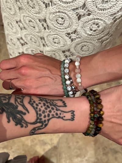 The image shows two arms resting together. The arm on the left is adorned with three beaded bracelets: a white and pink bead bracelet, a white and black bead bracelet with an evil eye charm, and a green bead bracelet. A ring is visible on the left hand. The arm on the right has two beaded bracelets in shades of green, brown, and gold, and a prominent tattoo featuring a stylized leopard figure. The background includes a textured white fabric on the left and an indistinct beige surface on the right. The image does not contain information about the location in the city of Puçol, Spain.