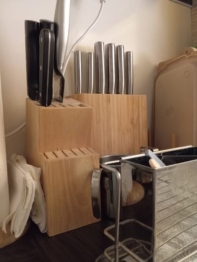 A close-up, low-angle shot depicts a kitchen counter with knife blocks, napkins, and a utensil holder. In the foreground, a stainless steel utensil holder contains several items, including what appears to be a blue object and a beige object, partially obscured by wire shelving. Behind the utensil holder, a wooden knife block is visible, with a stack of white napkins to its left. Further back, two wooden knife blocks are arranged, one with knives in slots and another with knives protruding from the top. A white electrical cord runs behind the knife blocks. To the right, a beige cutting board rests against a wall. The lighting is subdued, creating shadows on the counter. The image is taken indoors, likely in a residential kitchen. There are no people present in the image. The specific location within the city of Baden, Austria, cannot be determined from the visual information provided.