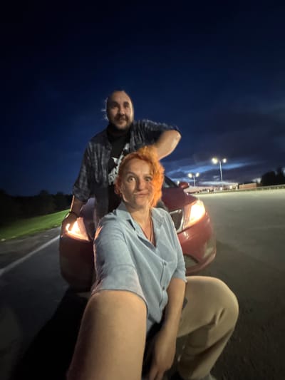 Two individuals, a man and a woman, are positioned in front of a red car at night. The man, with dark hair and a beard, is standing behind the woman. The woman, with bright orange hair, is in the foreground, facing the camera. They are in an outdoor setting, possibly a parking lot, with streetlights visible in the background and a dark sky above. The car's headlight is illuminated. The image appears to be a selfie. The location is identified as Alexeyevskoye, Russia.