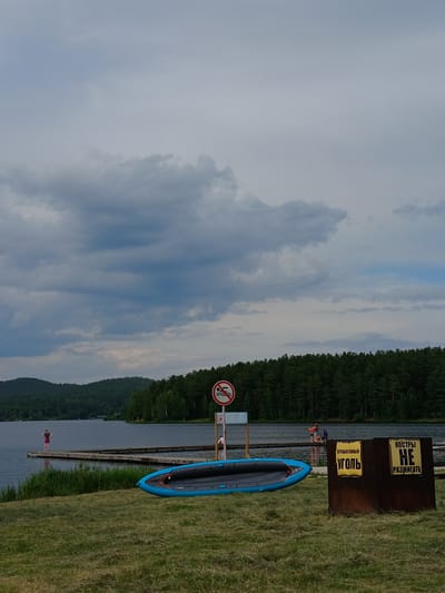 The image depicts a scene at a lakefront, likely in Revda, Russia, given the Cyrillic text on signs. In the foreground, a blue and black inflatable boat rests on grassy ground. To the right, two metal containers with yellow signs are visible. One sign reads "ОТРАБОТАННЫЙ УГОЛЬ" (Spent coal), and the other says "КОСТРЫ НЕ РАЗЖИГАТЬ" (Do not light fires). In the midground, a wooden pier extends into the calm lake, with several people standing and sitting on it. A person in pink shorts and a dark shirt stands on the far end of the pier. A warning sign with a red circle and diagonal line over a swimmer is posted near the pier. The background features a dense forest along the shoreline and rolling, tree-covered hills under a cloudy sky.