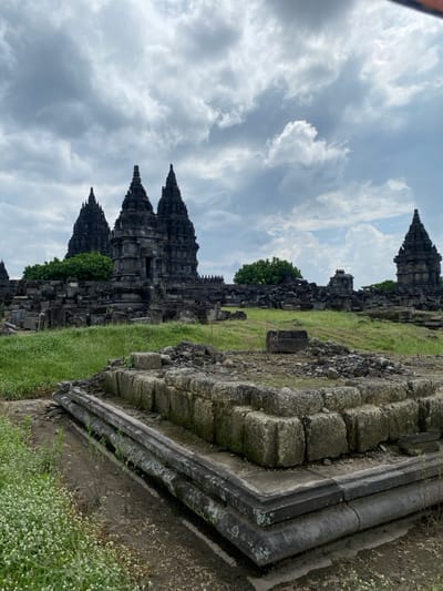 The image shows the ancient ruins of the Prambanan Temple Complex in Kecamatan Prambanan, Indonesia. The temple complex is composed of several stone temples in varying states of disrepair, including a large, three-tiered temple in the background. The foreground contains a large, square stone structure that appears to have once been a platform or base for another structure. The ground around the structures is covered in green grass, and some small trees can be seen growing in the area. The sky above is mostly cloudy, with some patches of blue sky visible. The photo was taken from a low angle, giving the viewer a perspective that emphasizes the size and scale of the ancient ruins.