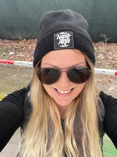 This image is a close-up, selfie-style photograph of an adult female located in the city of Oberhausen, Germany. The subject is centered, smiling directly at the camera with her teeth visible. She has long, blonde hair that flows over her shoulders and chest. She wears a dark, ribbed long-sleeved top and dark-tinted, aviator-style sunglasses with a thin metal frame. A dark, knitted beanie with a white square patch, featuring the stylized black text "NACHT AKTIV," covers her head. A portion of a dark-colored vest or backpack strap, possibly with a camouflage pattern, is visible over her right shoulder. The background is out of focus. Behind the subject, a dark green or gray solid barrier or wall is present. Below this barrier, the ground consists of bare earth with scattered dry leaves and small pieces of debris. A concrete curb with a distinctive red and white painted stripe runs horizontally across the mid-background. A paved surface is visible to the left of the curb and partially on the lower right. A small, bare plant stalk protrudes from the ground on the right side of the background. A segment of a bright green grated surface is visible at the bottom right corner of the frame.