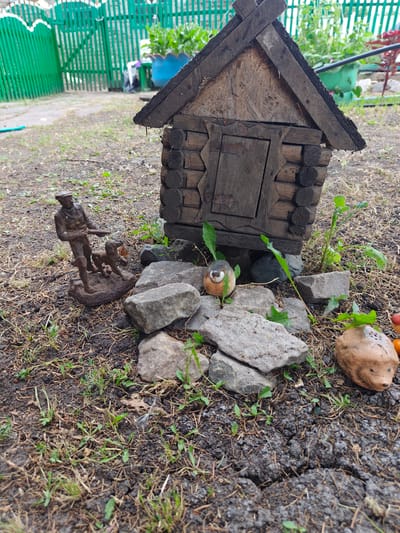 In a rural outdoor setting, possibly in Майкөл ауылдық округі, Kazakhstan, a small, rustic, log-built structure resembling a miniature house stands on the ground. In front of the house, a bronze-colored statuette depicts a man with a hat and a rifle, accompanied by a dog. Several rocks of various sizes are scattered around the scene. To the right of the house, a ceramic figurine of a hedgehog is visible. The ground is covered with dry grass, soil, and some small green plants. In the background, a green picket fence and lush vegetation are present.