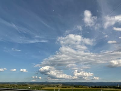 The image displays a vast expanse of the sky above a rural landscape. The sky is a vibrant blue, filled with a variety of clouds. Wispy cirrus clouds stretch across the upper portion, interspersed with larger, more voluminous cumulus clouds that cast subtle shadows. In the distance, a hazy mountain range forms the horizon, with rolling hills and fields of green and yellow stretching out below. A road or highway is visible in the foreground, with a guardrail along the bottom edge of the frame. Scattered streetlights and possibly utility poles dot the landscape. The overall scene depicts a bright, clear day in a pastoral setting, likely the outskirts of Orăștie, Romania, given the characteristic undulating terrain and agricultural fields. There are no people or discernible actions within the frame.