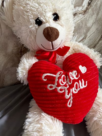 A plush white teddy bear is held by an unseen person. The bear has black button eyes, a brown nose, and a red ribbon tied around its neck. It is holding a large, red, heart-shaped pillow embroidered with the words "Love You" in white cursive letters. The background consists of a textured grey surface and a beige, patterned surface. The scene is indoors, likely in a domestic setting. There is no discernible location in Gagny, France.