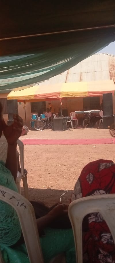 This image depicts an outdoor event taking place in Kasuwan Magani, Nigeria, under clear daylight. The perspective is from beneath a green overhead canopy, with two individuals partially visible in the immediate foreground, seated on white plastic chairs. The subject on the left wears a green patterned garment, with their arm and hand raised. The subject on the right wears a red fabric with white patterns.
In the midground, a temporary setup occupies an area of dry, light brown soil. A narrow, rectangular red carpet strip is laid directly on the ground, extending towards the background. Beyond this, a large temporary tent with yellow and orange fabric panels provides cover for a musical performance area. Under this tent, a blue drum kit is prominently positioned, flanked by two large, dark-colored PA speakers. An electronic keyboard is set up to the right of the drum kit. Multiple white plastic chairs are arranged within the tent area. Several individuals, identified as musicians, are present: one is seated behind the drum kit, another is at the keyboard, and a third is partially visible with an arm raised.
The background features a light-colored building with a corrugated metal roof. A barred window is visible on the building's wall, positioned behind the tent structure. The sky above the building and tents is clear and light blue.