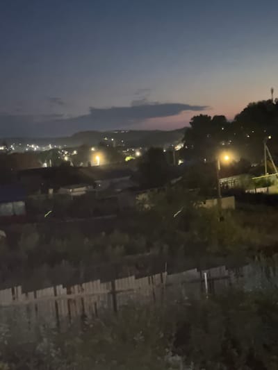 An outdoor, nighttime, low-light scene depicts a residential area, possibly in Mednogorsk, Russia, as suggested by the provided context, featuring a twilight sky with scattered clouds. The foreground is dominated by a weathered wooden fence and dense vegetation. In the midground, several houses with visible roofs and some illuminated windows are nestled amongst trees. Streetlights illuminate parts of the scene, casting a warm glow. In the background, a distant town or village is visible with numerous lights twinkling against the darkening landscape and hills. Two faint, horizontal green streaks are present in the midground, possibly indicating light trails or atmospheric phenomena. The overall impression is one of a quiet, rural or suburban settlement at dusk.