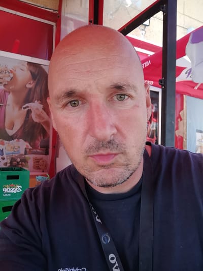 A selfie taken in Dralfa, Bulgaria shows a middle-aged bald man with light green eyes and a stubble beard. He is wearing a dark t-shirt with a lanyard around his neck. In the background, there is a red awning, a picture of a woman eating a dessert, and green crates. The lighting suggests it is daytime.