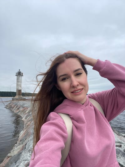 A woman with long, auburn hair stands on a stone jetty, holding her hand to her head. She is wearing a pink hoodie and a beige backpack. Behind her, a lighthouse stands on the jetty, with a body of water on either side. The sky is overcast. The location is identified as being in the city of Motornoe, Russia.