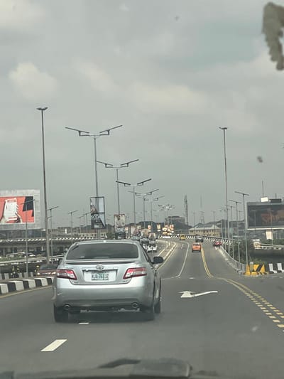 The image depicts an elevated multi-lane expressway in Ebute-Metta, Lagos, Nigeria, under a uniform overcast sky. The perspective is from within a moving vehicle, looking forward. A silver Toyota Camry (license plate KJA 761 JZ) is centrally positioned in the right-center lane, receding from the viewer. The road features white dashed and solid lane markings, double yellow dividing lines, and a large white right-turn arrow painted on the pavement. The bridge's safety barriers are marked with black and white stripes. Tall street light poles, some featuring multi-directional LED luminaires, line both sides of the expressway. In the mid-distance, an orange sedan and a red sedan are visible on the road. On a lower road segment to the left, beneath the elevated structure, two individuals on motorcycles are partially visible near a barrier. The urban background consists of various buildings and a prominent communication tower. Multiple large billboards displaying advertisements, including references to "SCENES Film City," are erected along the route.