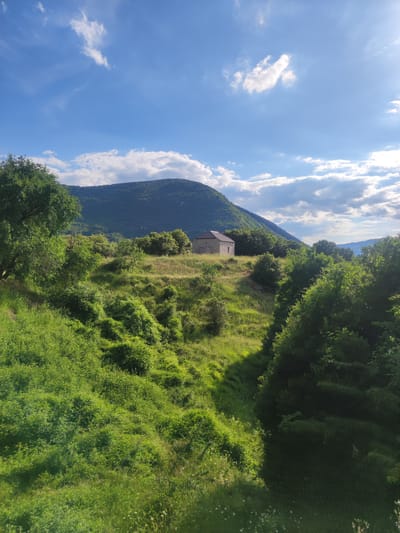 The image depicts a rustic stone building situated on a grassy hillside. The building is small and appears to be an old structure with a dark, sloped roof and visible stone walls. It is surrounded by lush green vegetation, including trees and bushes, creating a natural, somewhat overgrown landscape. In the background, a large, tree-covered mountain dominates the scene. The sky above is a vibrant blue, dotted with scattered white clouds, suggesting fair weather. The overall impression is one of a remote, tranquil rural setting. The image location is specified as Egnatia Municipal Unit, Greece.