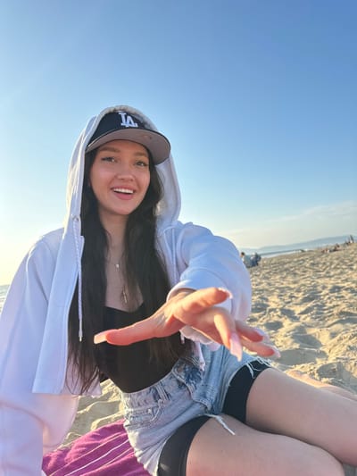 A female with long, dark hair, wearing a black Los Angeles Dodgers baseball cap, a white cropped zip-up hoodie, a black tank top, denim shorts over black undershorts, and a silver cross necklace, is depicted in a medium shot. She is seated on a fuchsia-colored towel on a sandy beach. Her right hand is extended forward towards the camera, fingers slightly spread, showcasing long, light pink acrylic nails. Her face is turned towards the camera with a wide smile, mouth slightly open. The background reveals a wide expanse of light-colored sand and the ocean in the distance under a clear blue sky. Several indistinct figures are visible further down the beach. The scene is illuminated by natural sunlight, suggesting an outdoor daytime setting at Manhattan Beach, United States.