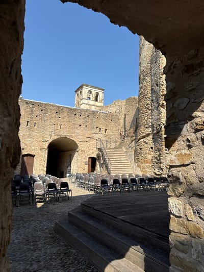 The image depicts the inner courtyard of a historic stone structure, likely a castle or ancient fortification, in Dolceacqua, Italy. The courtyard is set up for an event, with rows of empty chairs arranged on a cobblestone floor. In the foreground, a wooden stage with stairs is visible. The stone walls are weathered, with some showing signs of decay. A prominent arched entranceway leads into a dark interior. A stone tower, appearing to be part of the structure, stands against a clear blue sky. Sunlight casts distinct shadows across the courtyard.
