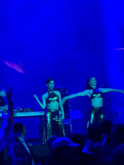 The image captures a dimly lit stage performance in Tsilkani, Georgia, dominated by a vibrant blue light. Two female performers are the focal point, both wearing shimmering, sequined outfits. The performer on the left has her hair styled up and is wearing a halter-neck crop top and flared pants. The performer on the right, also with her hair up, wears a similar halter-neck crop top and sequined pants, her arms extended outwards. A DJ setup is visible in the background, with turntables and mixers. In the foreground, the silhouettes of an audience are visible, suggesting a live event. The overall atmosphere appears energetic and theatrical.