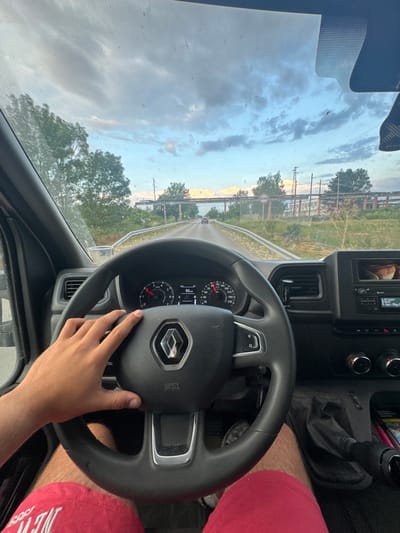 The image shows a first-person perspective from inside a vehicle, likely a van or SUV, driving on a paved road. A person's left hand is visible on the steering wheel. The steering wheel has a Renault logo in the center. The dashboard displays the speedometer reading 93 km/h. Outside the vehicle, the environment is rural with trees and vegetation lining the road. In the background, there is an industrial structure with overhead pipes and power lines. The sky is cloudy with patches of blue. The scene appears to be on the outskirts of Krushovitsa, Bulgaria, given the context of the request. The overall impression is of a journey in progress under a dynamic sky.