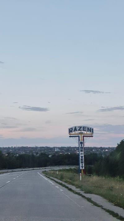 The image depicts a road sign at the entrance to Razeni, Moldova. The sign is tall and colorful, with the word "RAZENI" prominently displayed at the top. Below this, the number "1414" is visible, likely indicating the year the settlement was founded. Further down, the phrase "BINE ATI VENIT" (Welcome in Romanian) is written vertically. The sign is positioned on the right side of a paved road that curves to the left. In the background, a town with houses and church steeples is visible, nestled amongst trees. The sky is a pale blue with scattered clouds, suggesting it is either early morning or late evening. The overall scene is peaceful and evokes a sense of arrival.