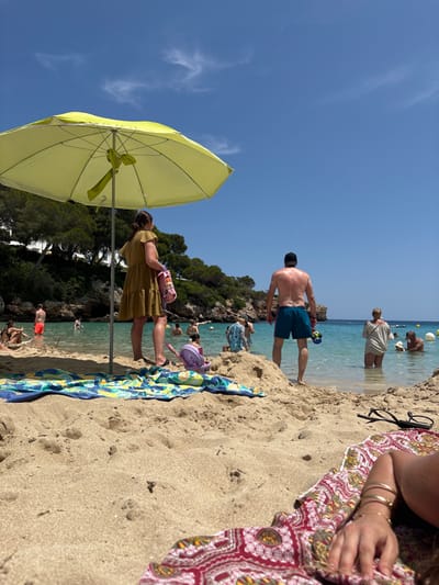 A sunny beach scene shows people enjoying the water. A man in blue swim trunks is partially submerged in the water. A woman in a khaki dress stands near a light yellow umbrella. Beachgoers are scattered along the shoreline, and a blue and green beach towel is laid out on the sand. Part of a patterned garment and a person's hands are visible in the foreground, suggesting the photograph was taken from a reclined position. Lush greenery surrounds the beach.