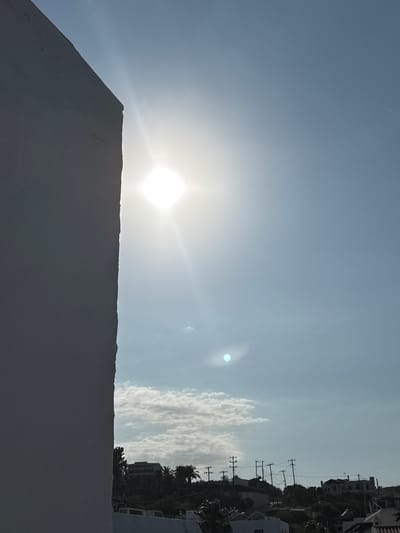 This media file captures a bright, sunny day in Chersonisos Municipal Unit, Greece. The sun is prominently featured, casting a strong glare across the sky and creating lens flares. The scene is dominated by a large, light-colored wall in the foreground, which takes up the bottom portion of the frame. Beyond the wall, the sky is a pale blue, with a few scattered, wispy clouds.

In the distance, on the right side of the image, a line of buildings and trees is visible, suggesting an urban or semi-urban environment. Utility poles with wires are also present, indicating infrastructure. The presence of buildings and vegetation hints at a developed area, possibly a residential or tourist district.

The lighting suggests it is daytime, likely mid-morning or afternoon, given the intensity of the sun. There are no people or active events depicted in the image. The overall mood conveyed is one of brightness and a clear, sunny atmosphere. No text is visible in the media file.