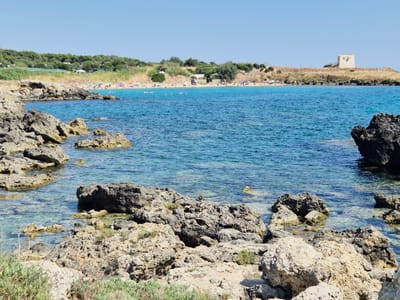 This image depicts a coastal scene in Leporano, Italy, featuring a rocky shoreline in the foreground with clear, turquoise water extending towards a sandy beach filled with people enjoying the sun and sea. The beach is lined with umbrellas and foliage, and in the background, a historic tower stands on a hill overlooking the coast. The sky is a clear, bright blue, indicating a sunny day.