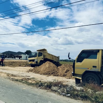 The image depicts a construction or land-clearing site under a bright, partially cloudy blue sky, crisscrossed by several overhead power lines. In the foreground, an asphalt road transitions into a dirt and gravel shoulder, with sparse green and dry vegetation, and some visible litter.

Two yellow dump trucks are prominent in the midground. The truck on the left is actively involved in earthmoving, with its dump bed raised, having deposited a large pile of light brown soil onto the site. A person, dressed in a blue and red jacket, stands near the front left side of this truck, observing or working. The front of this truck displays a license plate with the alphanumeric sequence "BG 9042 ZK".

To the right, the front cabin and wheel of a second yellow dump truck are partially visible, appearing to be parked or waiting on an elevated dirt mound, facing towards the left.

In the background, several modern houses with dark roofs are visible amidst a developing landscape. Beyond these structures, a flat, open area with faint green vegetation stretches towards the horizon. The scene suggests ongoing construction or groundwork during the daytime in Keudealuerheung, Indonesia, as indicated by the license plate.