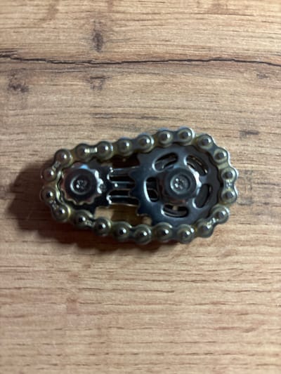 Centered against a wood-grained surface is a metal object. It appears to be a decorative piece constructed from a bicycle chain and two sprockets. The chain forms an oval outline, encapsulating the sprockets. The sprockets are different sizes, connected by a small metallic piece. The item is silver in color.