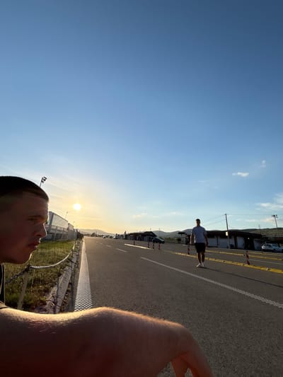 The image shows a road in Dimitrovgrad Municipality, Serbia, at sunset. A man in a gray shirt and black shorts is standing in the middle of the road. The sun is setting in the distance. The image is taken from a low angle, with the photographer's arm visible in the foreground.