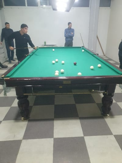 A billiards game is underway with several people in the room. One individual is focused on a shot, holding a cue stick. Another person stands aside, arms crossed, observing the game. A third individual appears to be watching as well. The billiard table is green and has several white balls and one red ball on its surface. The floor is tiled in a checkerboard pattern.