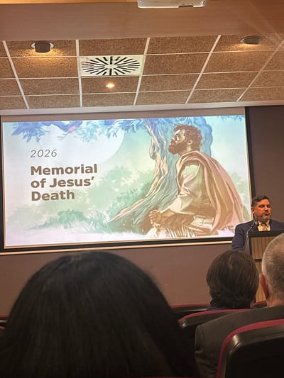 A slightly rotated image taken indoors, likely in an auditorium or conference room. A presenter stands in front of a projection screen displaying artwork depicting a biblical scene, presumably related to Jesus. The screen has the words "2026 Memorial of Jesus' Death" displayed prominently. The presenter, a man with dark hair and a beard, is wearing a suit jacket and speaks into a microphone. In the foreground, portions of attendees' heads and shoulders are visible, suggesting a seated audience. The room's wall features a grid-like pattern with ventilation systems and lighting fixtures. The location is specified as Sant Vicent del Raspeig, Spain, though this detail isn't evident from the image itself but provided as context.