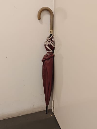 An umbrella with a curved wooden handle and a dark red fabric is leaning against a white wall in the corner of a room. The umbrella is closed and secured with white straps. The floor is dark gray and the wall has some minor scuff marks. The scene appears to be indoors. There are no people present. The image does not provide enough information to determine its location in the city of Settimo Torinese, Italy.