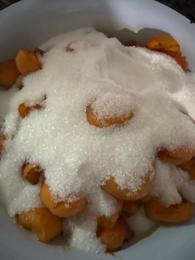 The image shows a close-up, overhead view of halved apricots covered in granulated sugar in a white bowl. The apricots are bright orange and appear to be fresh. The sugar is piled high, obscuring some of the fruit. The scene is likely located in an indoor setting, possibly a kitchen. There is no information in the image that would indicate a specific location in the city of Большие Озерки, Russia.