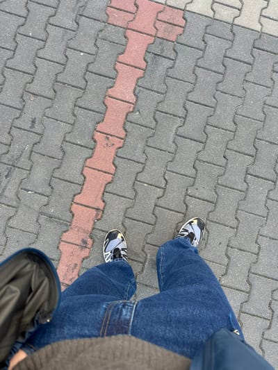 An overhead, first-person perspective photograph captured in Wolica, Poland, depicts the lower torso and feet of an individual standing on a paved surface. The ground is composed of interlocking "H"-shaped concrete pavers, predominantly dark grey, with a single diagonal stripe of reddish-brown pavers extending from the upper left to the lower right. The individual wears loose-fitting blue denim jeans and athletic shoes featuring black, grey, white, and yellow accents. A portion of a brown or olive garment is visible at the bottom center and left, and a dark blue or black garment with a metallic zipper is visible at the bottom right. Minor discoloration and dirt are present on some of the pavers, particularly within the reddish-brown stripe. No other individuals or significant objects are visible within the frame.