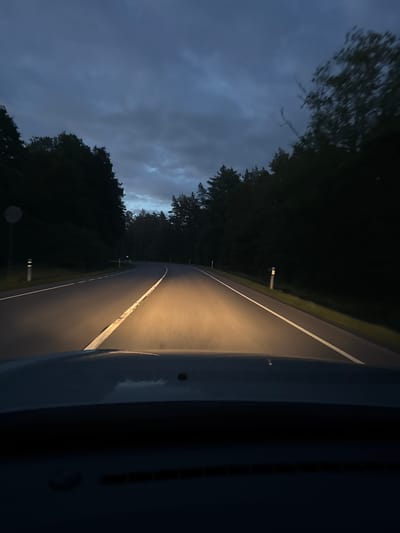 The image shows a view from the driver's seat of a car at dusk, driving on a paved road surrounded by a forest. The headlights of the car illuminate the road ahead, which curves to the left. The sky is dark blue with heavy clouds. There are white road markings and reflective bollards on the side of the road. The dashboard of the car is visible at the bottom of the frame. The location is identified as Kloogaranna, Estonia. There are no people or other vehicles visible in the image. The scene appears to be quiet and isolated.