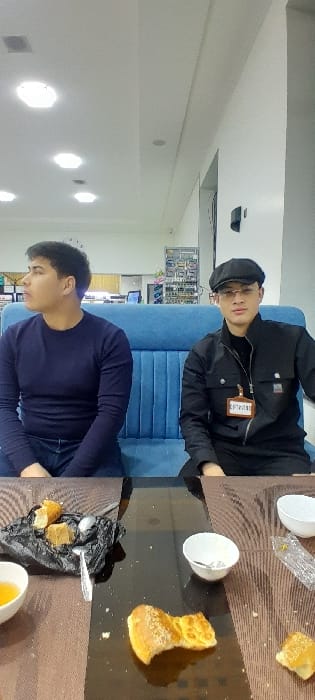 Two men sit on a blue sofa by a glass table. The man on top wears a black jacket and hat with glasses. He has a name tag. Below him is a man in a blue shirt. The table has a brown mat with a snack plate on the lower right. It also has white bowls and pieces of bread scattered around. A black plastic bag is seen with food and a spoon on the table.