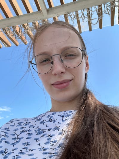 A young woman with long brown hair and glasses is looking up at the camera. She is wearing a white dress with a blue floral pattern. The background is a clear blue sky with some wooden structures and lights above her. The image appears to be taken outdoors in a sunny location, possibly a patio or pergola.