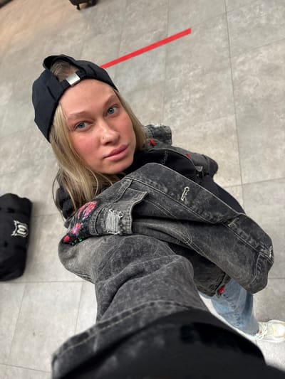 A young adult woman is taking a selfie in what appears to be an indoor space with light grey tiled flooring. The woman is wearing a black baseball cap, a black Calvin Klein t-shirt, a faded black denim jacket with patches, and light blue jeans. She has blonde hair and light eyes. A black bag is partially visible on the floor near her. A red stripe is marked on the floor. The city of Boldino, Russia is not identifiable within the image.