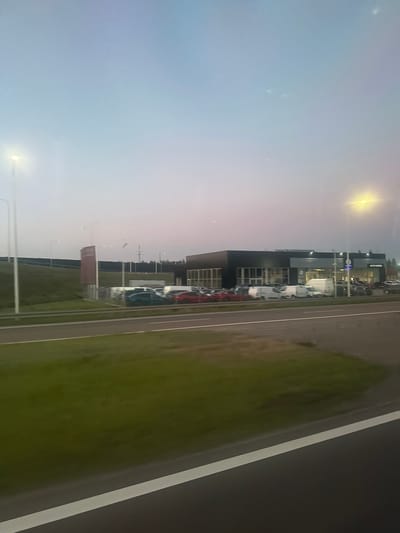 The image captures a twilight scene of a modern car dealership, identified as "AUTO-RAIDA" in Świdnik, Poland, along the express road DK17. The dealership features a dark, contemporary facade with large glass windows, displaying a selection of vehicles parked in front and on the lot. Among the vehicles are several cars, including what appears to be a blue hatchback and red sedans, alongside white vans.
The dealership is situated on a slight incline, with a grassy embankment to its left. The background includes a dark fence line and a cluster of trees, suggesting a semi-rural or developing urban outskirts setting. Several tall streetlights are visible, illuminating the area as dusk settles. One prominent sign, partially obscured, is a vertical red rectangular structure with a logo. The foreground shows a portion of the DK17 road, with a white line marking the lane. The sky is a gradient of pale blue and pink, indicative of either dawn or dusk. No people are clearly discernible in the image.