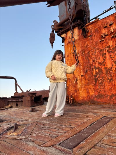 A young girl stands on the deck of a rusted shipwreck in Teriberka, Russia. She is wearing a light yellow jacket and grey sweatpants. The ship's hull, a vibrant orange with heavy rust, towers behind her, adorned with a thick metal chain. The deck is made of weathered wooden planks covered in sand. The sky is a clear, pale blue. The scene suggests a desolate, industrial coastal environment.