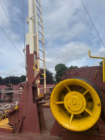 The image depicts the deck of a vessel in Brunsbüttel, Germany. The primary focus is on a large, yellow-painted metal wheel, possibly a winch drum or a capstan, situated on a dark red painted deck. The wheel has a visible serial number, "SS 020", stamped into its surface. To the left of the wheel, a substantial red metal structure supports a chain and a yellow lever mechanism. A white pole, possibly a mast or a ladder support, is positioned further left and has a metal ladder attached to it. Ropes are coiled on the deck in the foreground and midground. In the background, dark red ship structures and some trees are visible under a cloudy sky. The scene appears to be static, with no people or activity evident.
