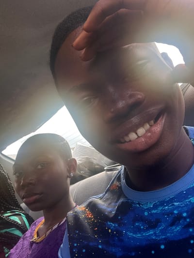 The image depicts two individuals within the interior of a vehicle in Eneka, Nigeria. The foreground subject, positioned to the right, is a dark-complexioned individual with short, dark hair, wearing a blue t-shirt featuring a multi-colored, cosmic-like pattern. This individual is smiling, revealing upper teeth, and has their right hand partially obscuring their forehead and upper facial area. A prominent lens flare originates from the upper left, crossing the foreground subject's face.
In the background, to the left, a second dark-complexioned individual is visible, characterized by closely cropped dark hair, a purple top with a lace-like texture, and a small, round, silver-colored stud earring in the left ear. This individual maintains a neutral expression. Both individuals are oriented towards the camera. The vehicle's interior includes dark-toned seats and a light-colored overhead section. A bright, rectangular window is visible in the upper background, and light-colored bags, possibly luggage, are stacked behind the background individual.