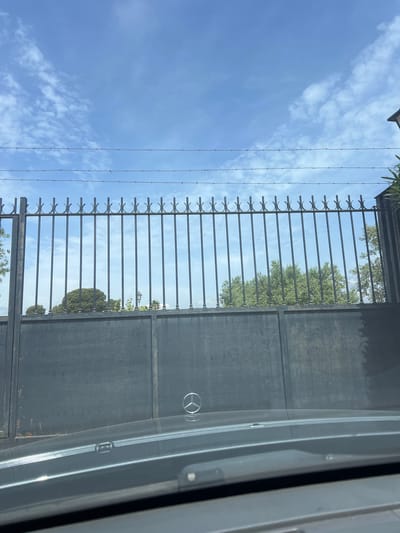 The image captures an outdoor scene, seemingly taken from inside a vehicle, looking out through a window at a tall, dark gate and the sky beyond. The perspective suggests a slightly upward gaze.
In the foreground, a distinct reflection of a Mercedes-Benz steering wheel logo is visible in the lower right portion of the image, superimposed on the gate. This indicates the photograph was taken from within a car. On the far right, parts of the car's interior, possibly a window frame or door panel, are visible.
The main subject is a substantial dark metal gate or fence. The lower section of the gate appears solid. Above this, there's a panel composed of vertical metal bars, each featuring a decorative, C-shaped or semi-circular element at its top. Further above this section, several strands of barbed wire are stretched horizontally, acting as an additional security feature.
In the background, a bright blue sky dominates the upper part of the frame, adorned with scattered white, wispy clouds. Through the openings in the gate's railings and above its structure, numerous green trees with dense foliage are visible. Some leafy vegetation also peeks into the frame from the top left corner, possibly belonging to a nearby building or overgrown plants.
The scene is brightly lit, indicating it is daytime with sunny weather. There is no human activity depicted, contributing to a static and calm atmosphere. The image provides a glimpse of a secured property boundary in Vila-seca, Spain.