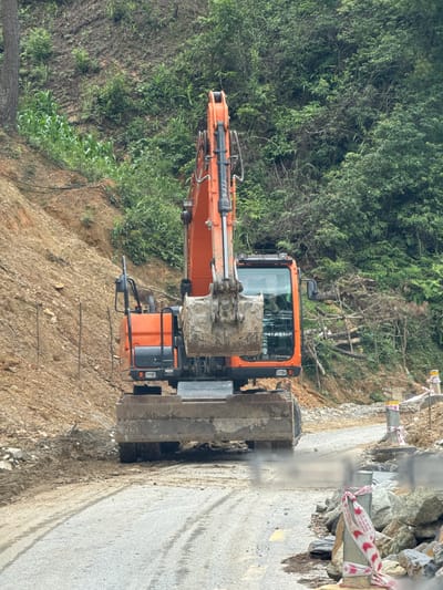 This image, captured in Pung Luong, Vietnam, displays a rural road with an orange and black excavator positioned centrally. The excavator faces forward, resting on a worn asphalt road surface marked by tire tracks and a faint yellow center line. The front blade of the excavator is lowered, and its bucket is visible, coated in dirt. A subtle haze, possibly exhaust or dust, emanates from beneath the machine.

To the left of the road, a steep, reddish-brown dirt hillside rises, partially covered with sparse green vegetation, including rows of young corn plants. A thick tree trunk is visible on the far left. Several slender wooden stakes are inserted vertically into the slope, and dark cables or hoses run horizontally near the base of the hill.

To the right of the road, another steep hillside is covered in dense green foliage, characteristic of a forested or jungle environment. Piles of cut logs and branches are visible against this slope. Along the right edge of the road, rocks and construction debris are present, alongside two white poles wrapped with red and white striped warning tape, indicating a construction or hazard zone. No individuals are visible in the scene. The overall environment suggests ongoing road construction or maintenance work in a mountainous or hilly area.